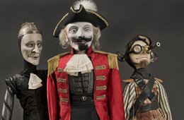 Freiherr von Münchhausen und seine Gefährten Johann und Berthold, Handstockpuppen von Matthias Hänsel mittig ein Mann in Uniform, neben ihm zwei futuristisch wirkende Personen