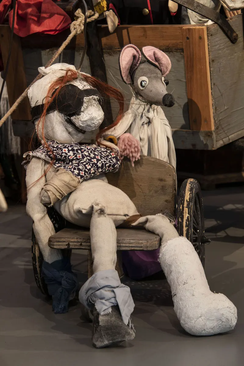 Eine Puppe mit Augenbinde und Gips am Bein, neben der eine Maus steht