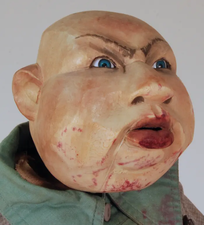 Männlicher Puppenkopf mit rot verschmierten Lippen und strengem Gesichtsausdruck.