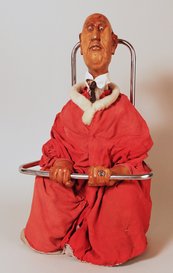Puppe: sitzender, glatzköpfiger Mann in großem Mantel